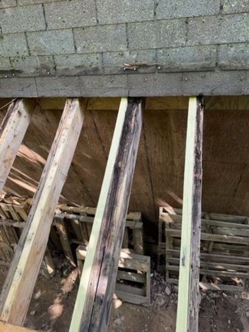 Damaged wooden deck framing beneath a stone wall, with exposed beams and debris underneath