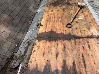 Damaged plywood roofing with a shovel on top beside a shingled roof edge