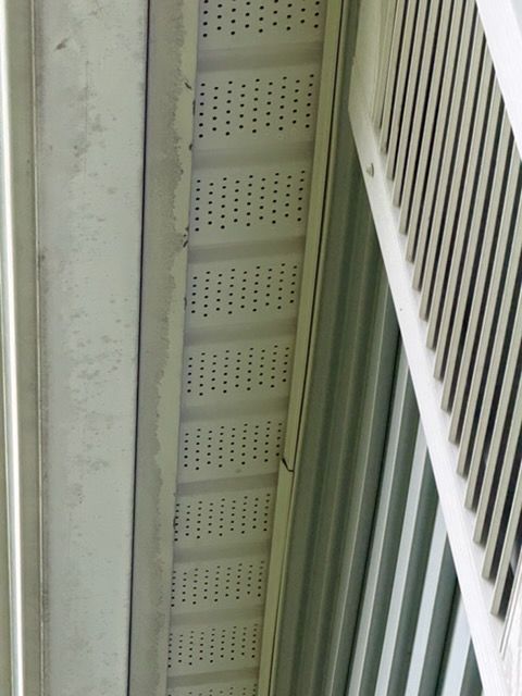 Close-up of a building exterior with white siding, perforated soffit vents, and a vertical metal pipe.