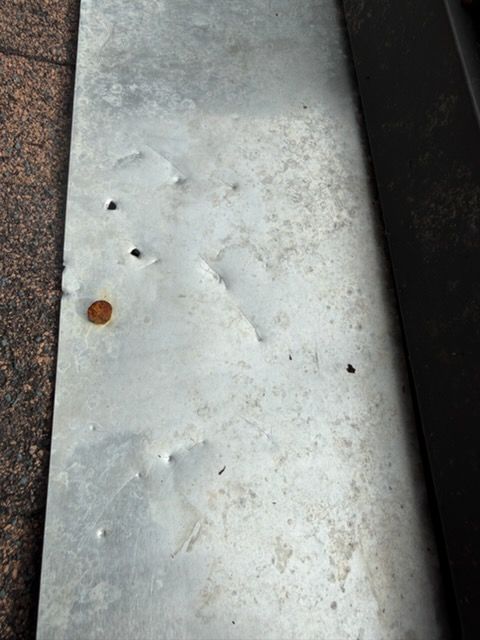 Close-up of a light gray metal surface with small holes, dents, and a rusty spot рядом dark edge