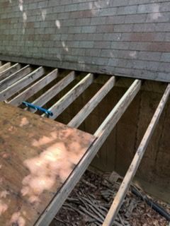 Wooden deck framing under construction beside a gray brick wall, with exposed joists and dirt below