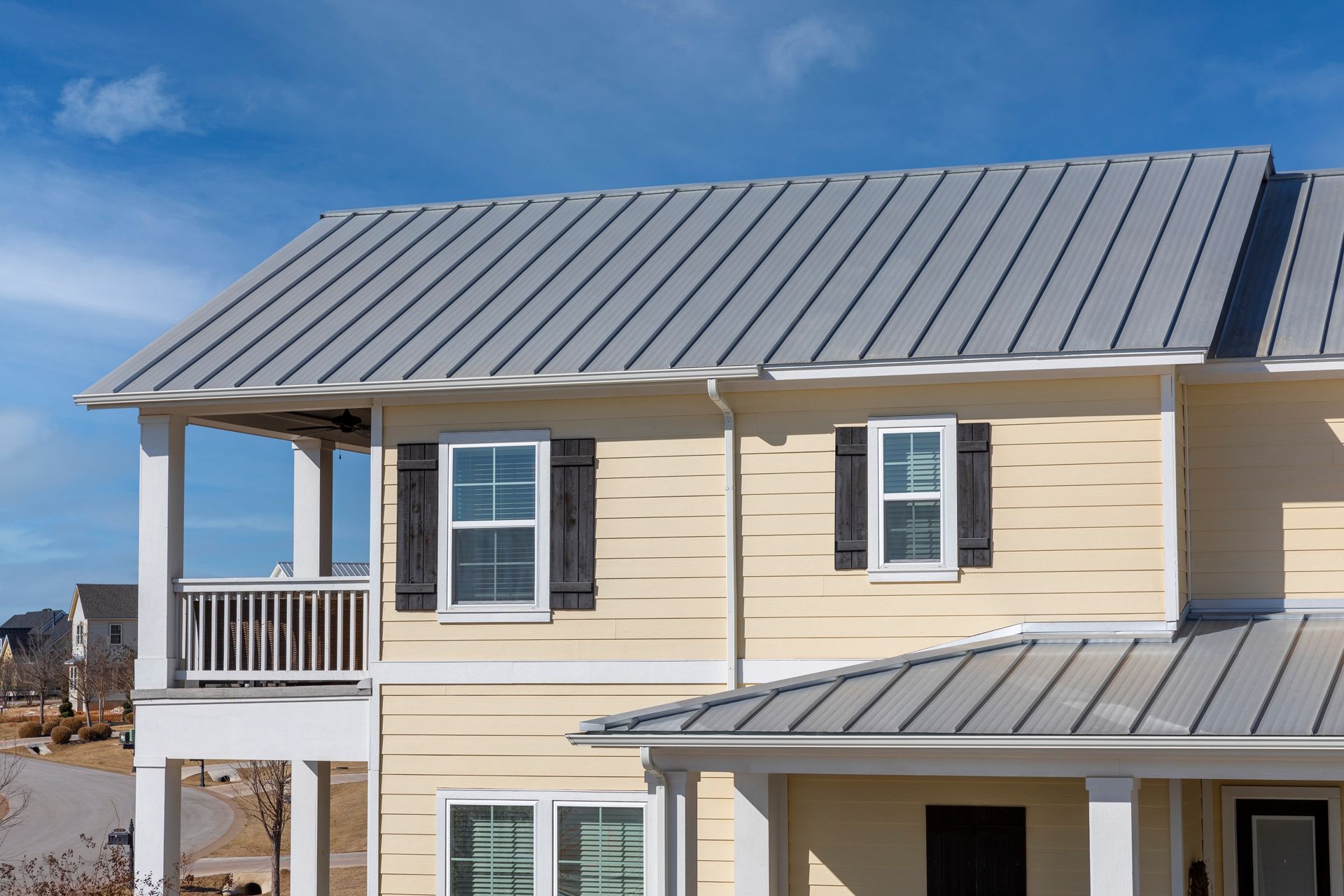 Two-story house with light yellow siding, gray metal roof, and blue sky background.