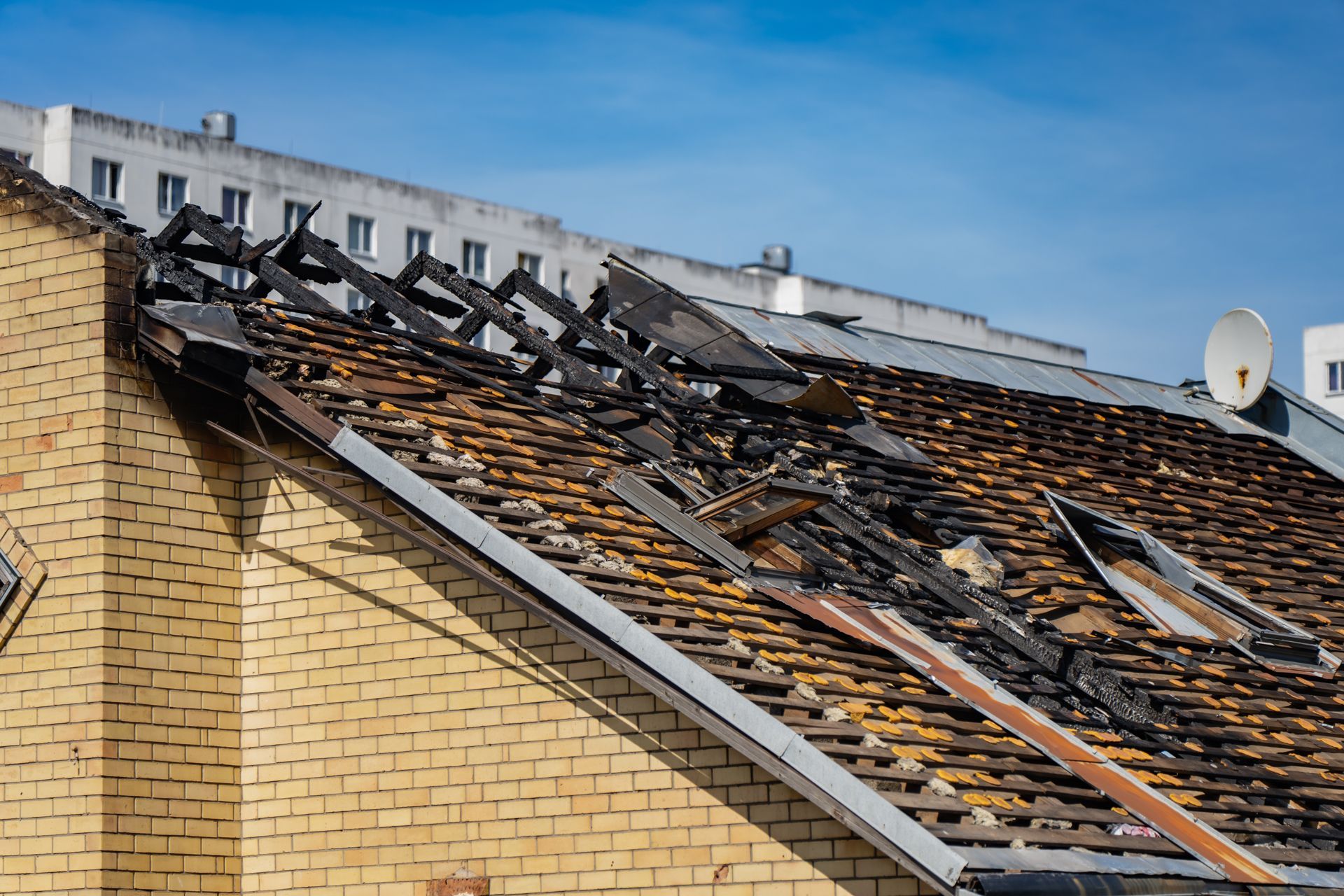 Damaged rooftop with missing shingles and exposed beams on a brick building under a blue sky