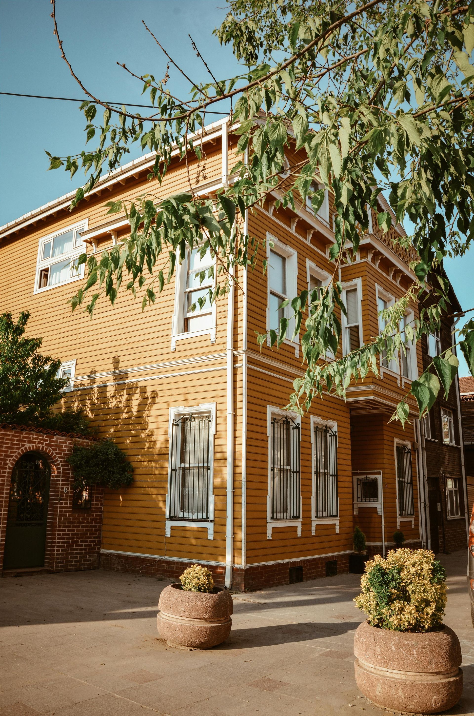 Yellow wooden house with white trim, green trees, and flower pots in a sunlit outdoor setting.