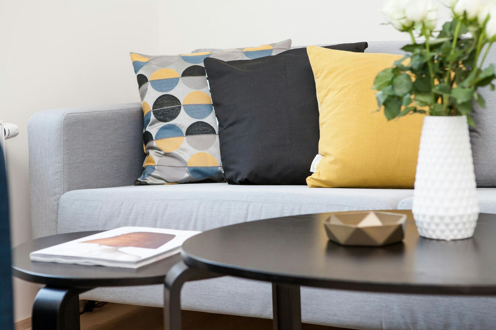 Gray couch with pillows (yellow, black, patterned) and two black coffee tables, a vase of flowers.
