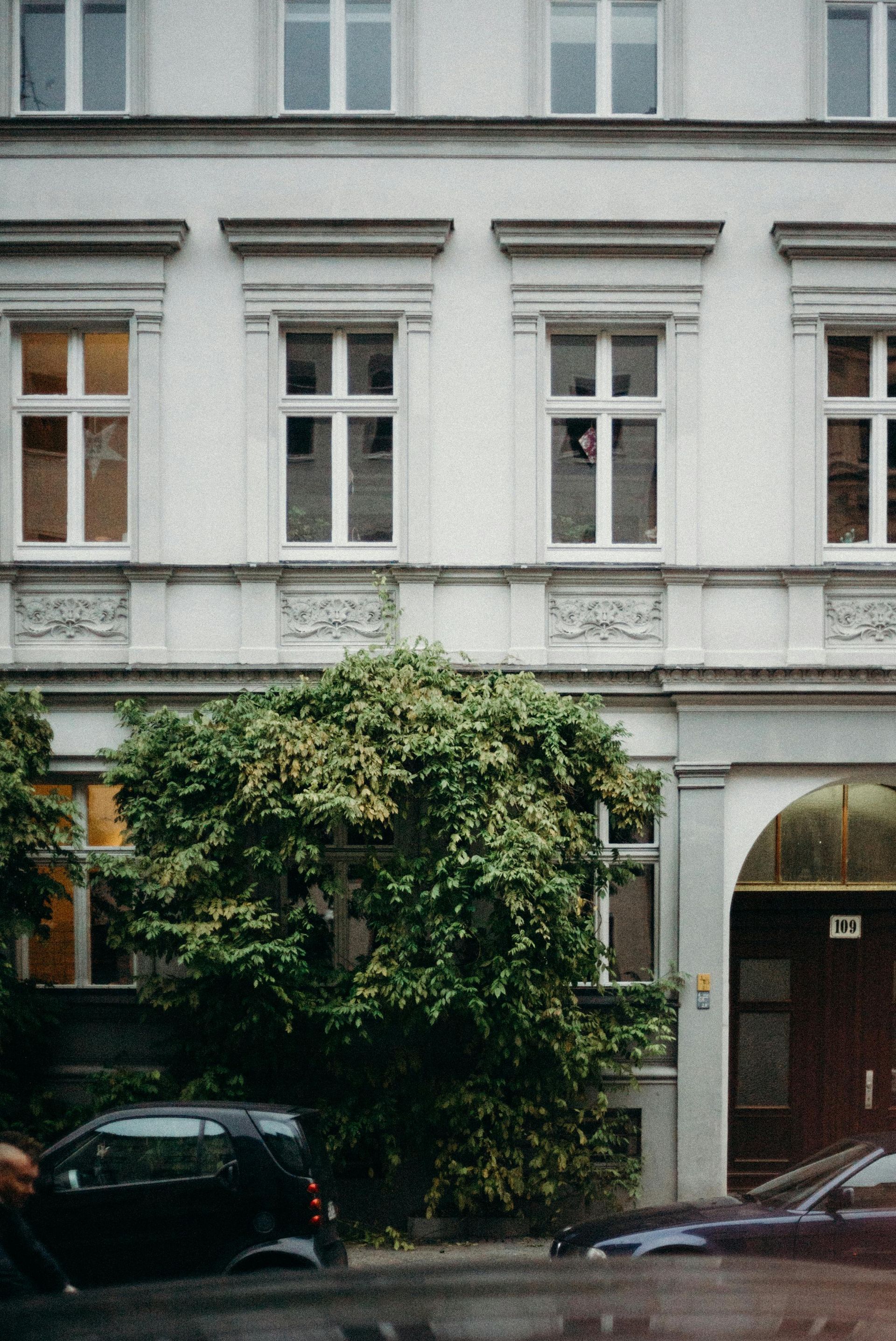 Building facade with windows, ornate details, and a green bush. A car is parked on the street.