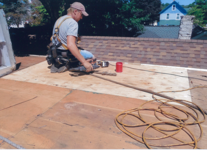 Roofer on a roof, wearing a safety harness, working near plywood. Brown shingles in the background.