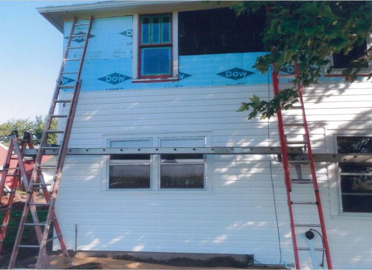 House exterior under renovation with ladders and scaffolding. Blue wrap covers the upper portion, white siding below.