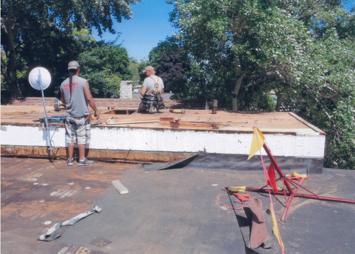 Two people working on a flat roof. One stands, the other kneels. Trees and a white building are in the background.