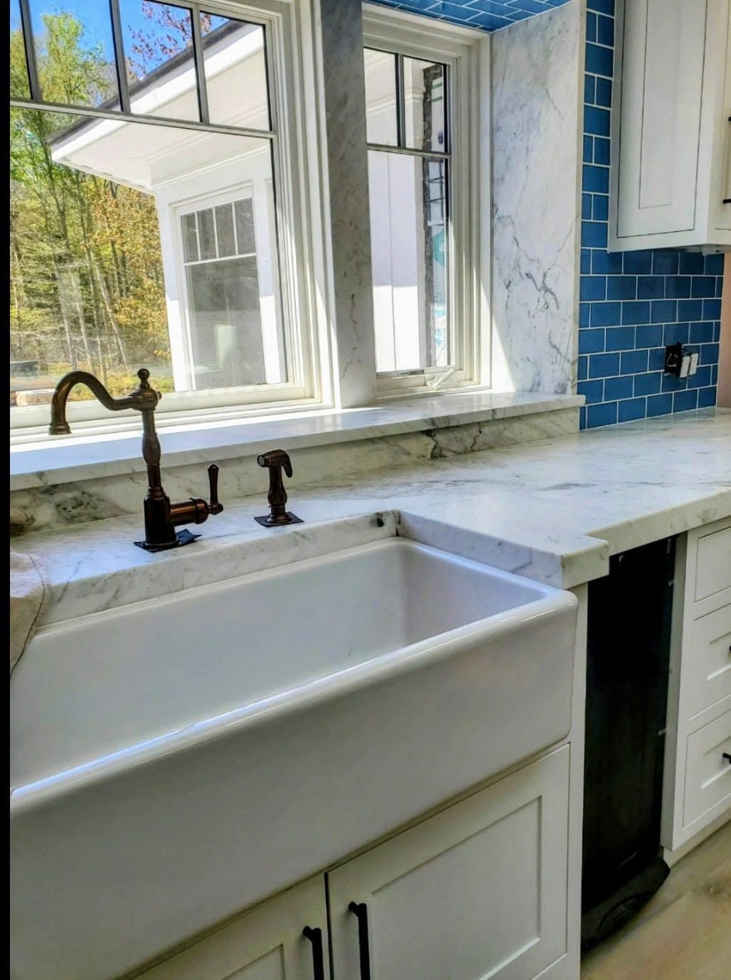 White farmhouse sink and brass faucet in a bright kitchen with blue tile backsplash and open window.