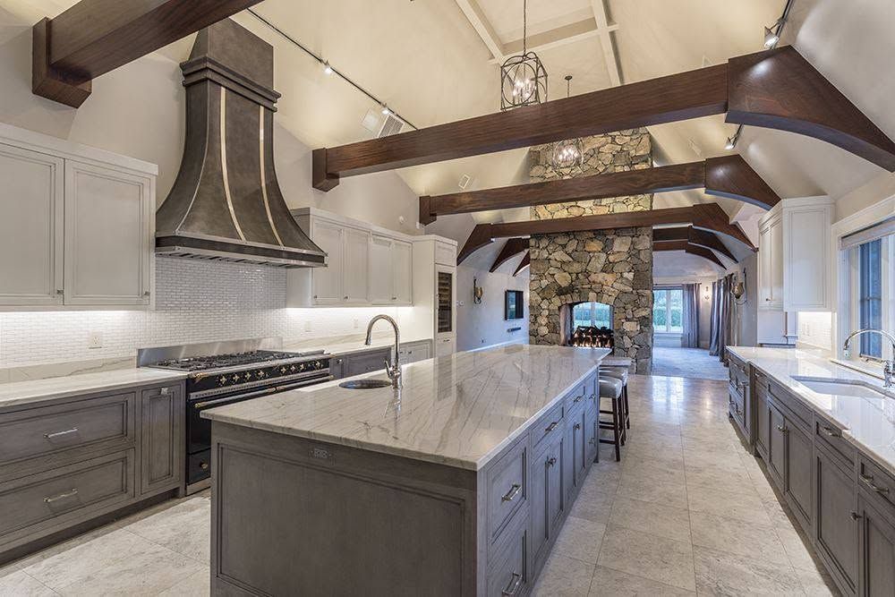 Modern kitchen with large island, marble counters, white cabinets, exposed beams, and stone fireplace at the far end