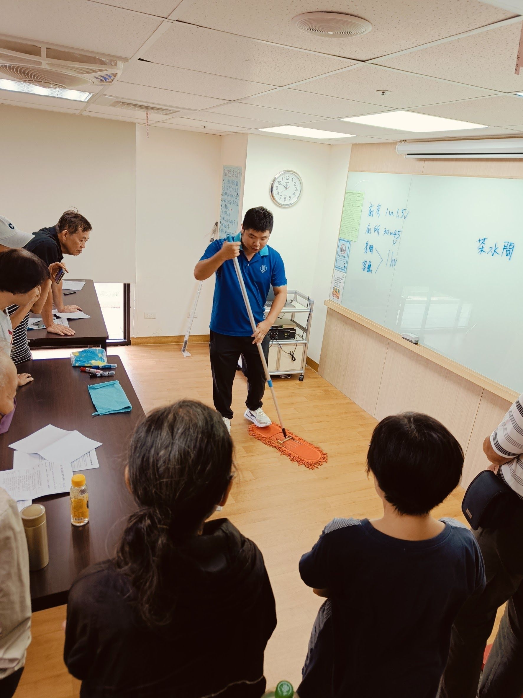 一名男子在教室裡用拖把示範,學生們在一旁觀看。背景中有一塊白板。