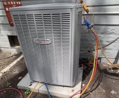 An outdoor air conditioning unit with gauges connected, on a concrete pad next to a wall.