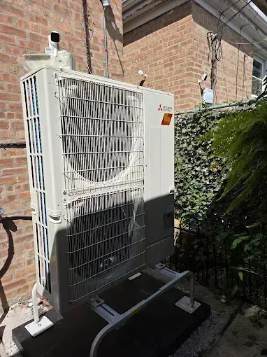Outdoor Mitsubishi heat pump unit next to a brick wall and foliage.