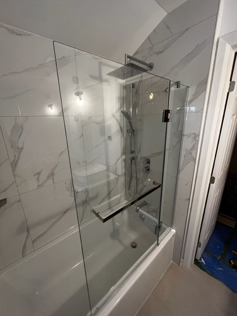 Une douche fermée par une paroi de verre transparent avec porte coulissante, baignoire blanche intégrée, pomme de douche mura