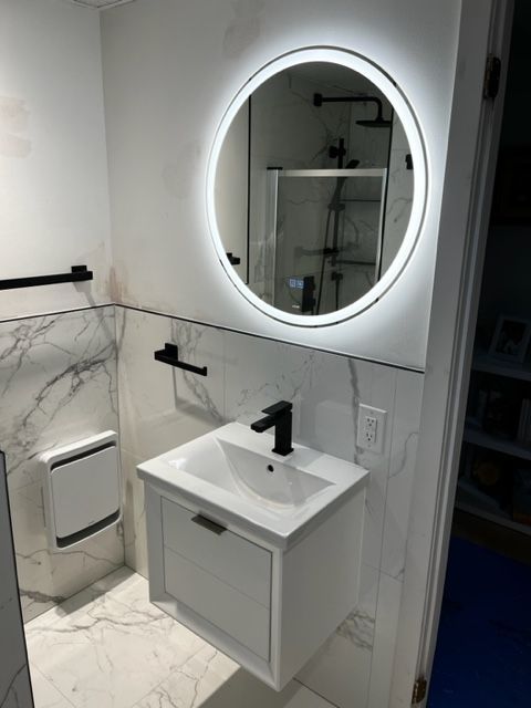 Salle de bain moderne avec miroir ovale illuminé, lavabo blanc, robinetterie noire, carrelage blanc et gris, et douche vitrée