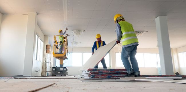 Des ouvriers du bâtiment installent un revêtement de sol dans une pièce lumineuse en cours de finition, avec des outils et des matériaux éparpillés sur le sol.