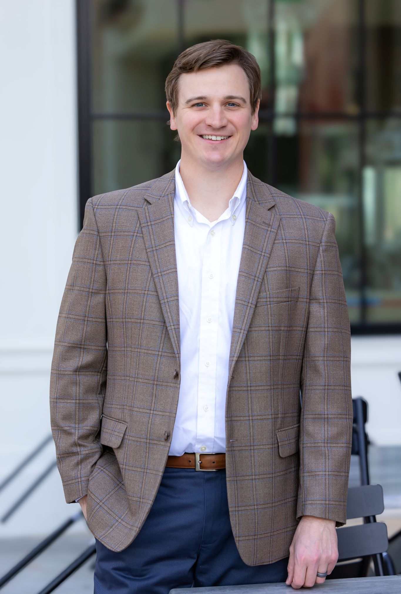 Man standing outdoors in a brown blazer and white shirt, smiling in front of a building.
