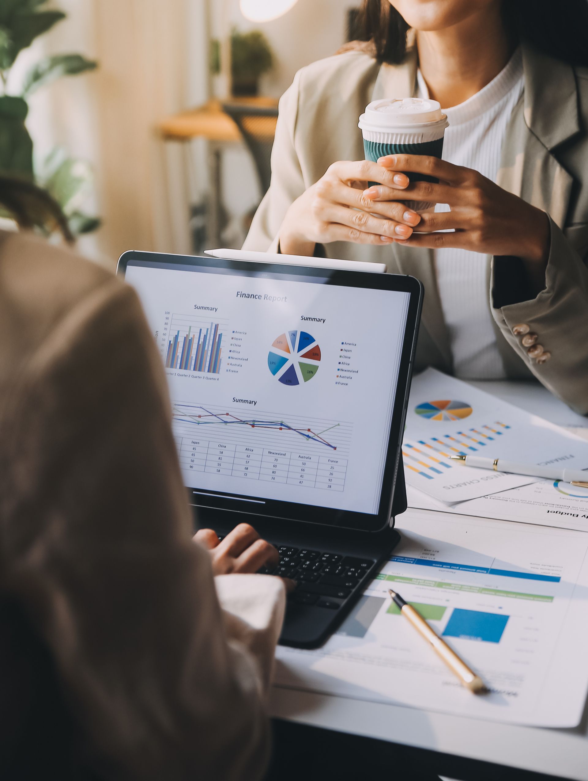 Two people at a table, analyzing charts and data on a tablet, while one holds a coffee.