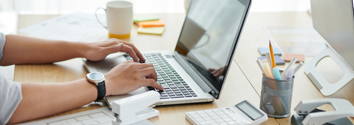 Hands typing on a laptop at a tidy desk with a coffee mug, stapler, calculator, and pen holder.