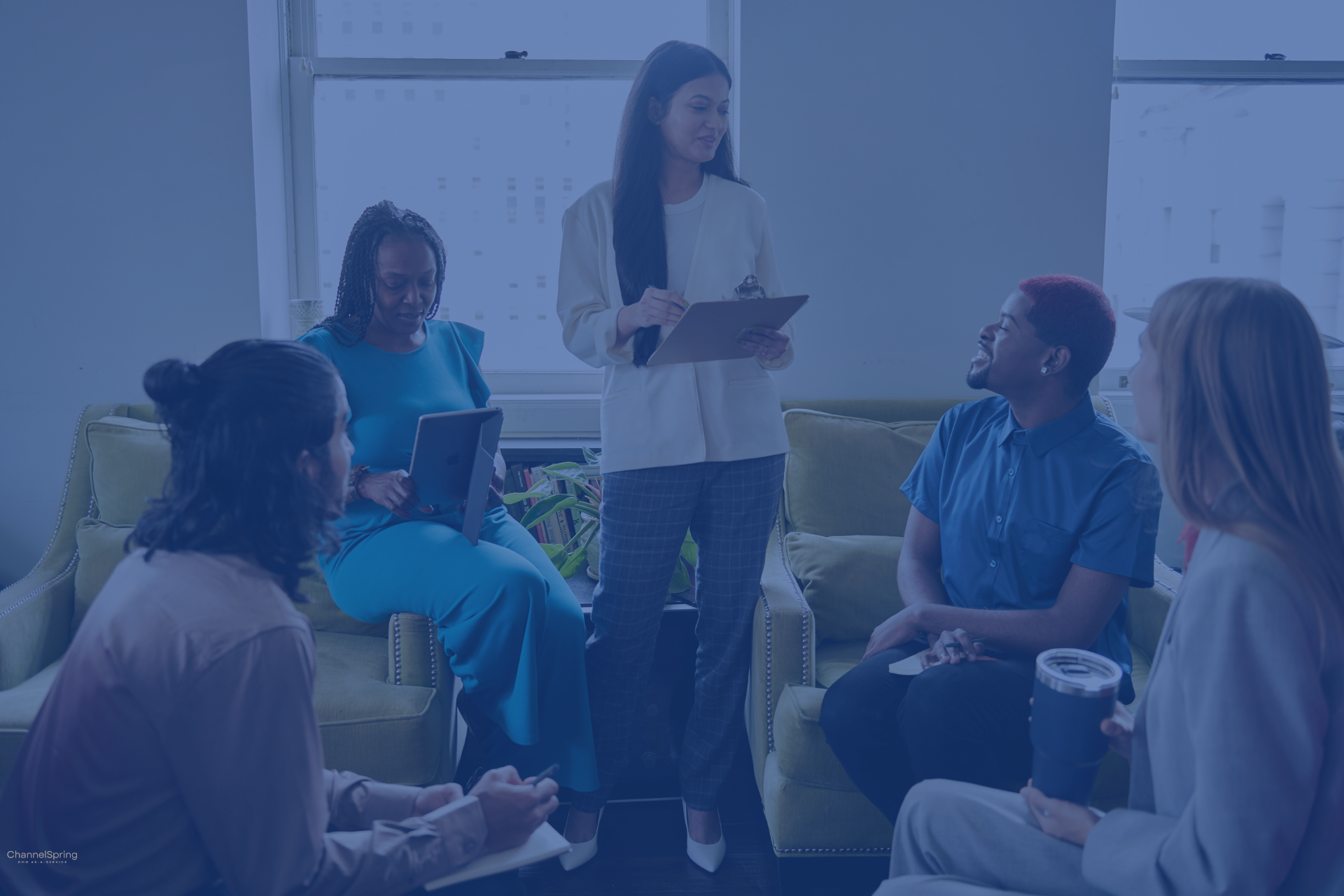 A marketing leader with a clipboard leads a planning session with a seated team in an office lounge.