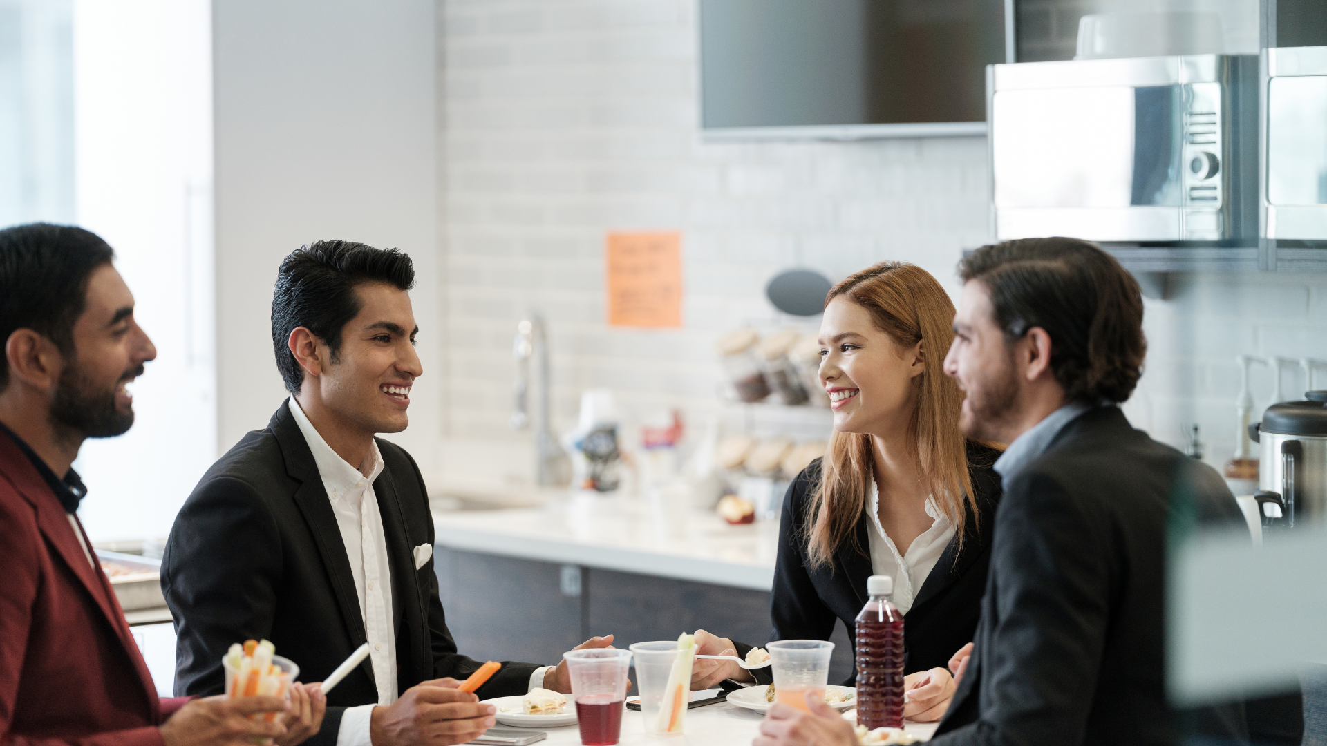 Four colleagues in business attire laughing together at a breakroom table, eating lunch.