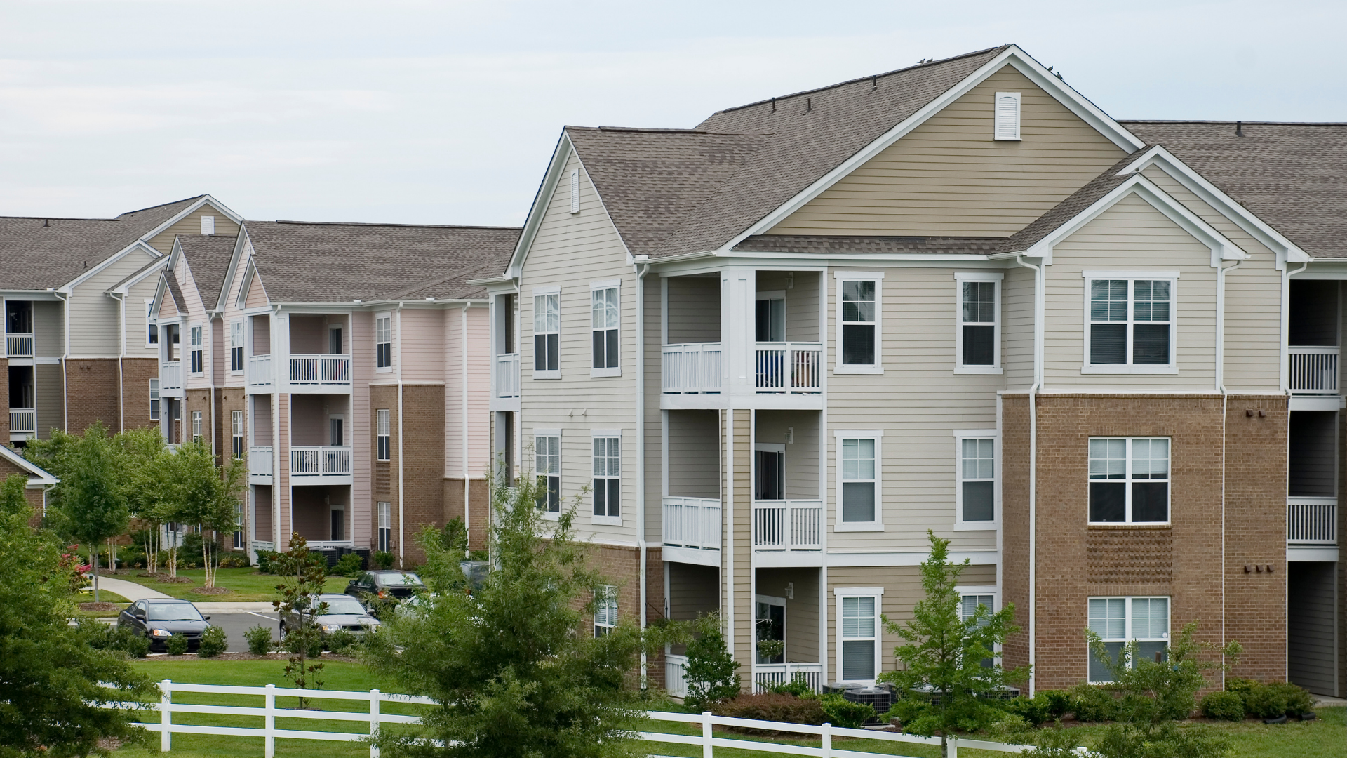 Apartment complex with beige and brick siding, balconies, and brown roofs under a cloudy sky.