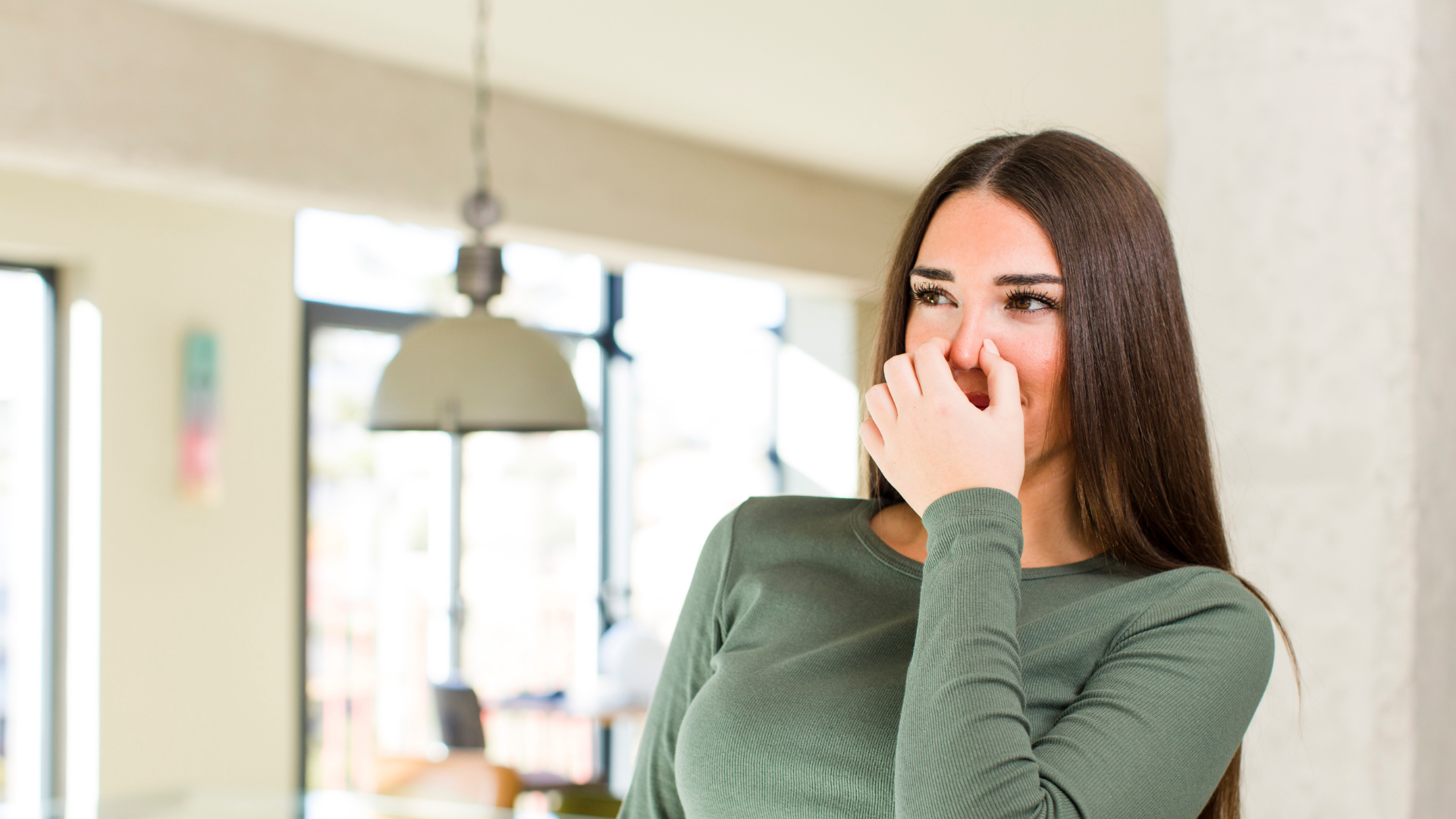 Woman in a green top covering her nose indoors, with a blurred kitchen in the background.