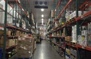 A person operates a forklift in a long, narrow aisle of a warehouse lined with tall shelves stacked with inventory.