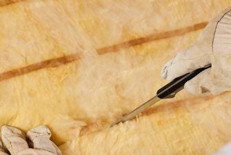A gloved hand uses a utility knife to cut a yellow fiberglass insulation batt.
