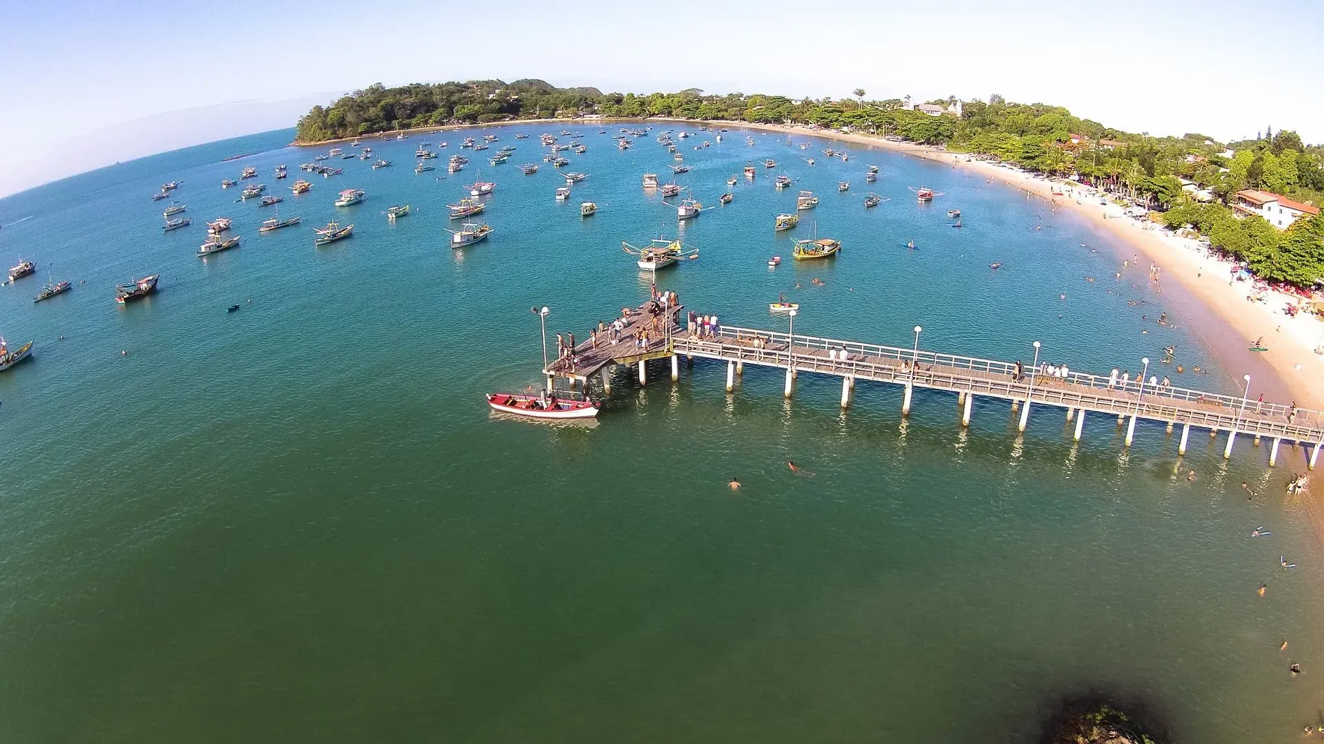 Vista aérea de uma praia com um píer se estendendo em direção ao oceano; barcos estão espalhados na água e pessoas estão no píer e na praia.