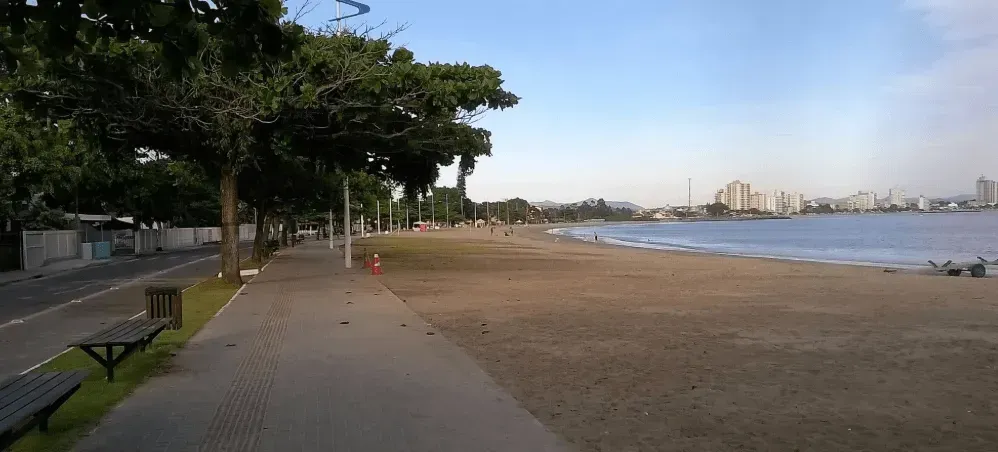 Uma cena de praia com um calçadão, árvores e o horizonte da cidade ao fundo. A areia é bege e a água é azul.