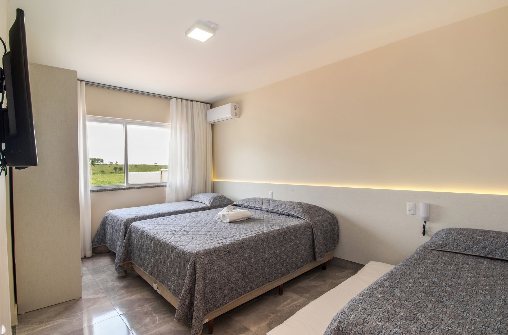 Um quarto de hotel com três camas, uma janela e uma TV na parede. Paredes bege e cabeceira branca.