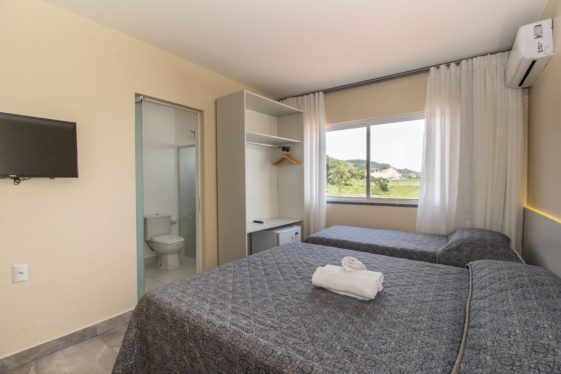 Quarto com duas camas, banheiro pequeno, TV na parede e janela com cortinas claras.