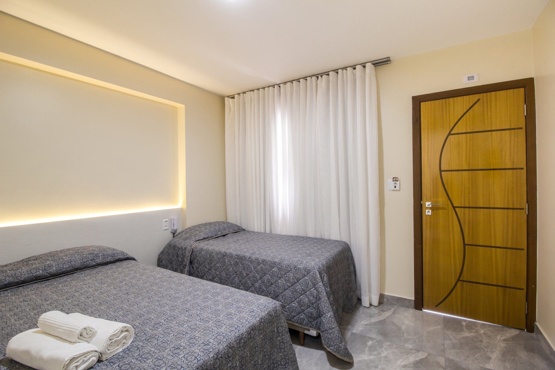 Duas camas de solteiro em um quarto com paredes bege. Uma porta à direita e uma janela com cortinas brancas no meio.