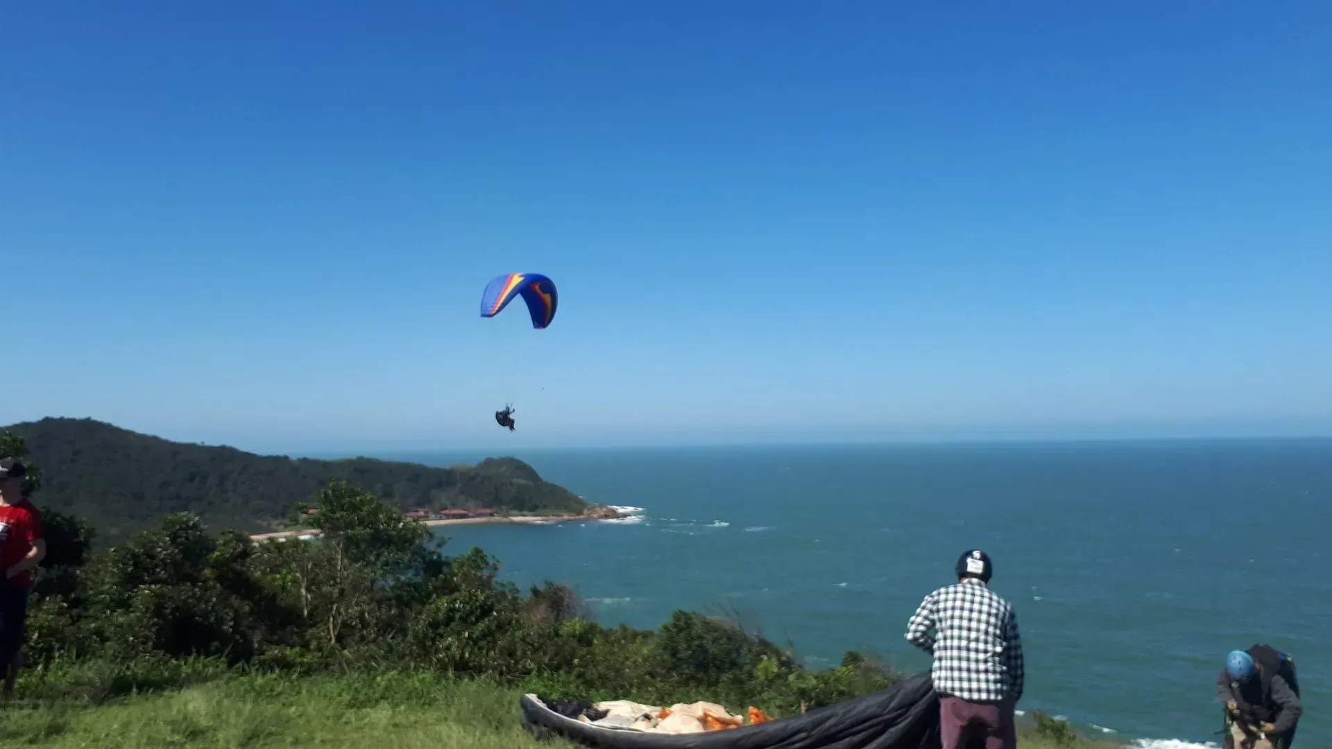 Dois parapentes sobrevoando o oceano, lançados de uma colina gramada com espectadores. Céu e água azuis brilhantes, litoral verdejante.