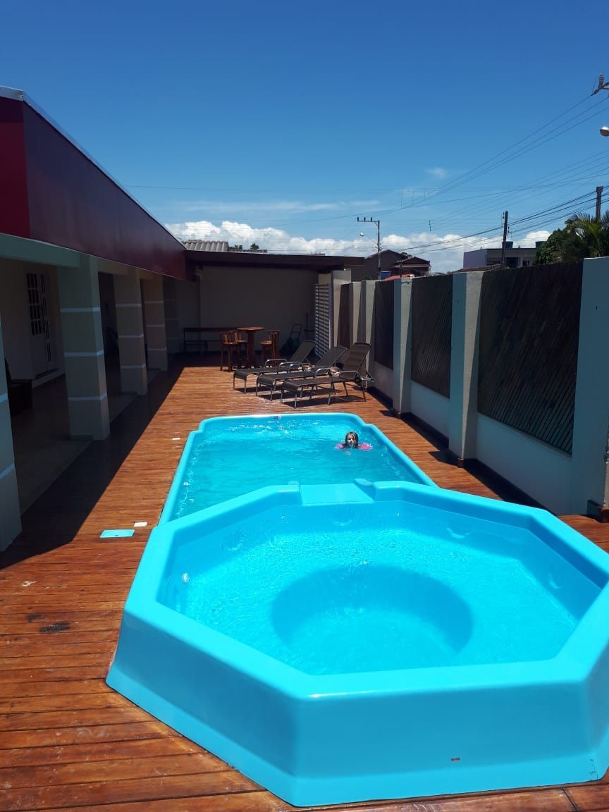 Uma piscina hexagonal azul e uma piscina retangular sobre um deck de madeira sob um céu azul brilhante. Uma pessoa nada na piscina retangular.