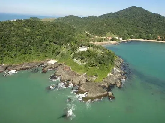 Vista aérea de uma península verdejante encontrando um oceano azul-petróleo. Uma pequena estrutura branca ergue-se no ponto, cercada por vegetação.