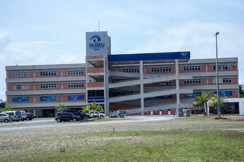 Vista externa do edifício MUSBU, uma estrutura de vários andares com rampa, em uma área gramada com carros estacionados. O céu está nublado.