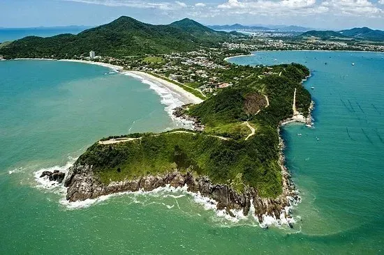 Vista aérea de uma península verdejante projetando-se em águas azul-turquesa, com uma praia e uma cidade visíveis ao fundo.
