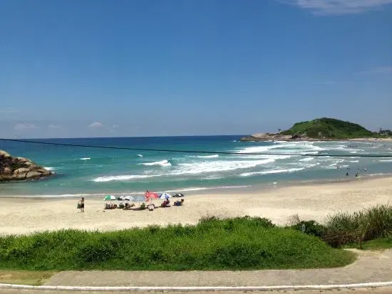 Cena de praia com céu azul, água turquesa e areia branca. Grama verde em primeiro plano, pessoas e uma pequena ilha ao fundo.