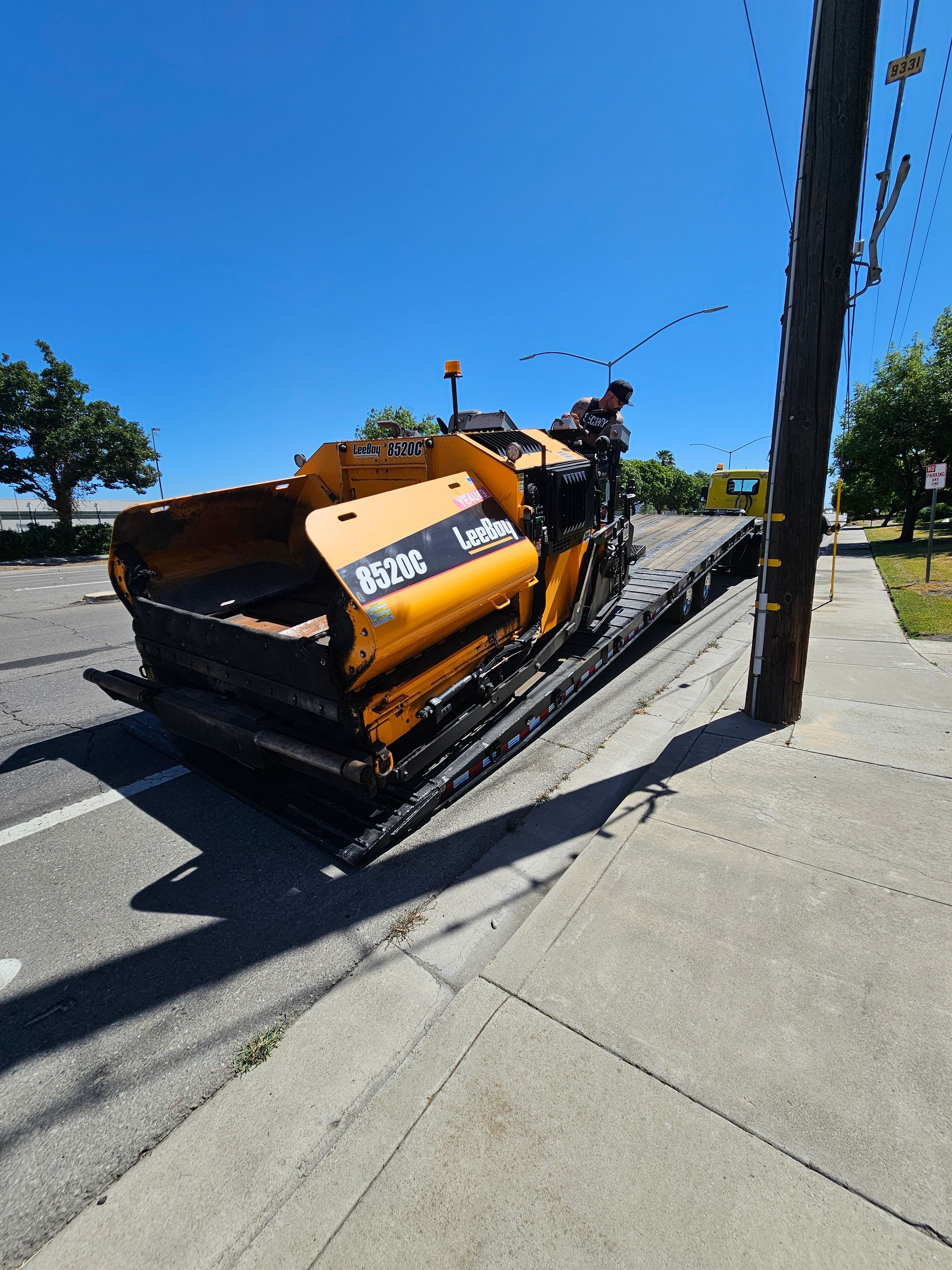 A yellow asphalt paver is being loaded onto a flatbed trailer on a sunny day next to a utility pole.