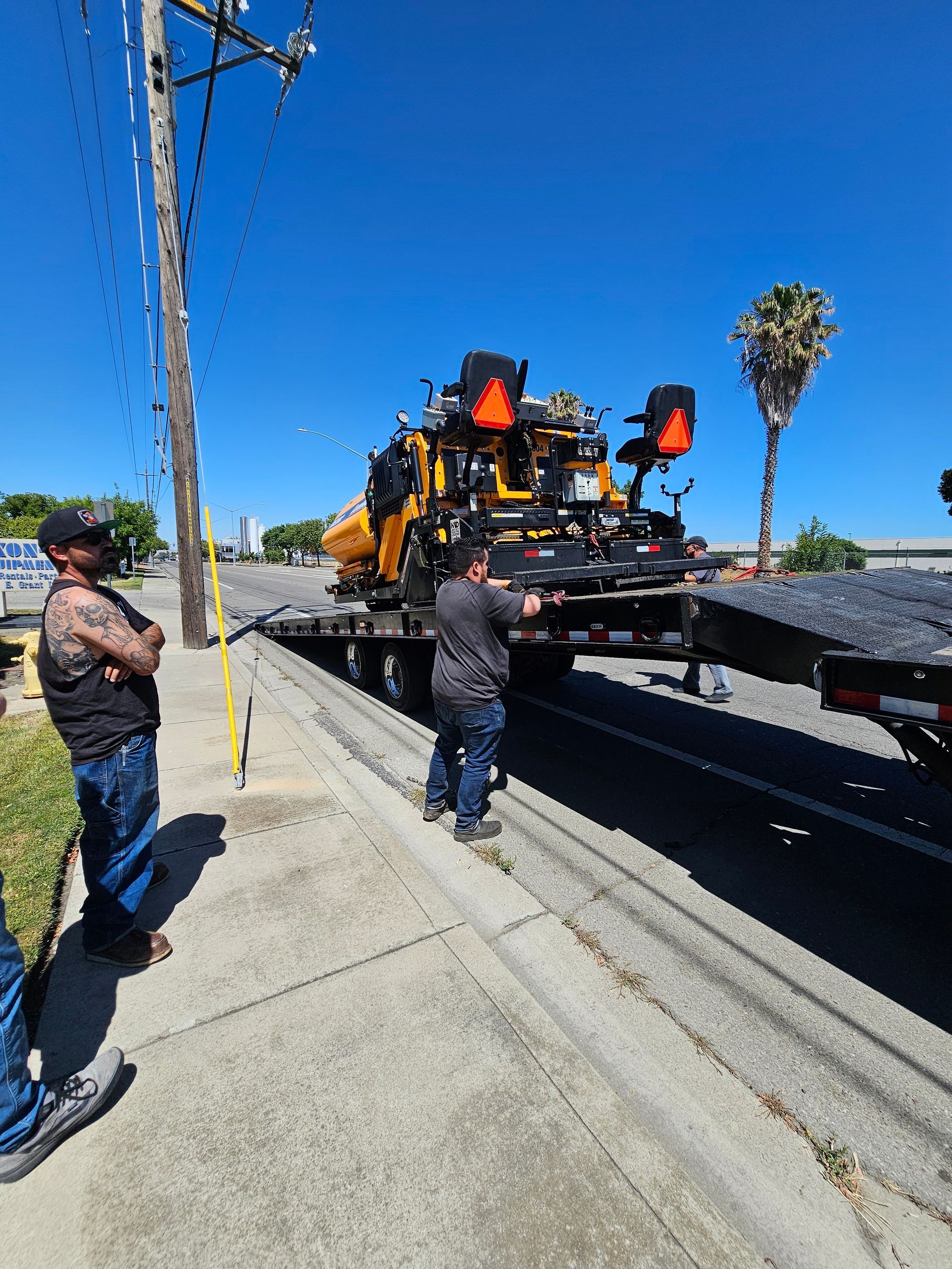 Two workers standing on a sidewalk next to a yellow construction vehicle being loaded onto a flatbed trailer.
