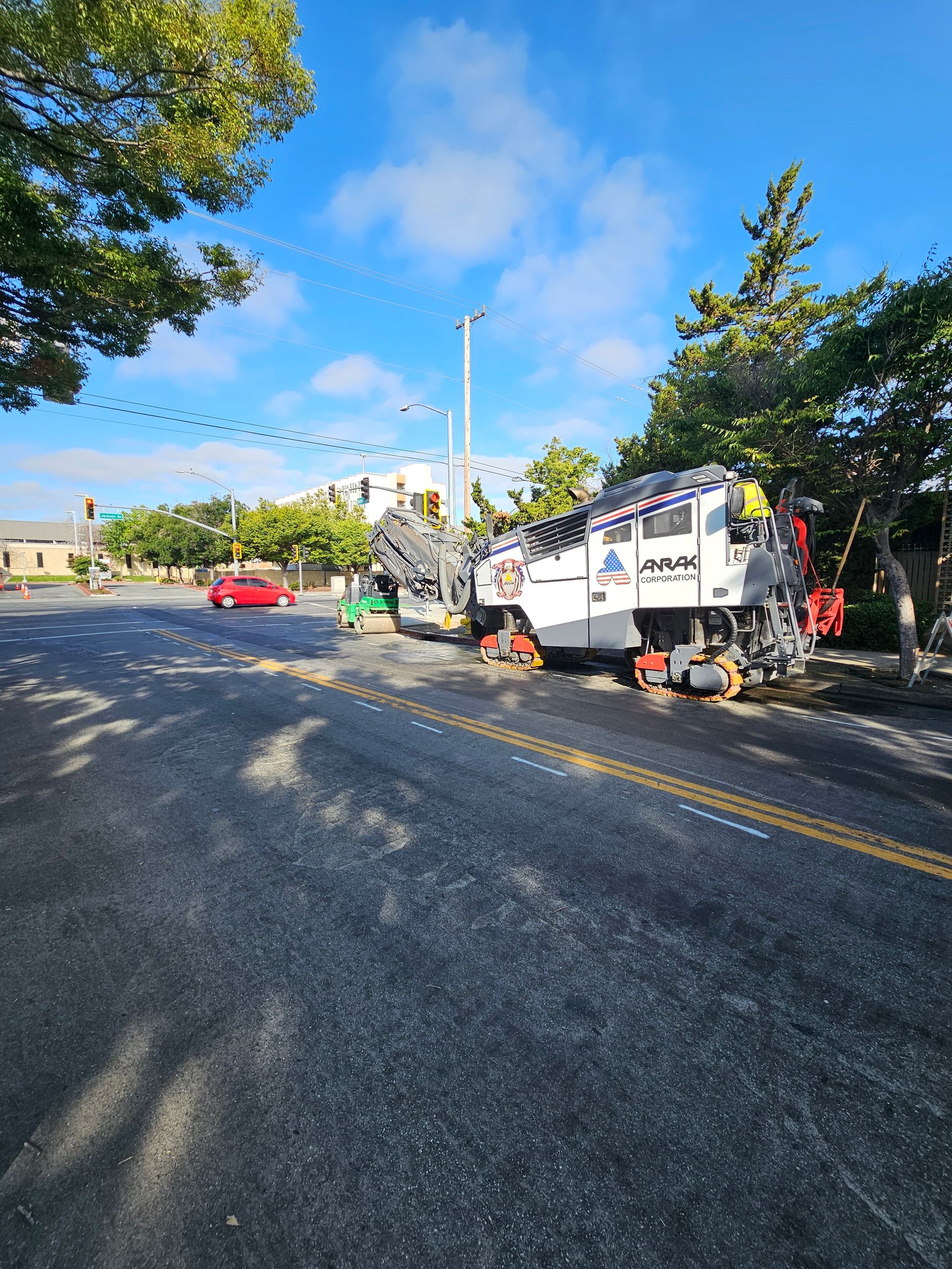 A large white construction vehicle parks on the side of a paved road near trees, with a red car visible in the distance.