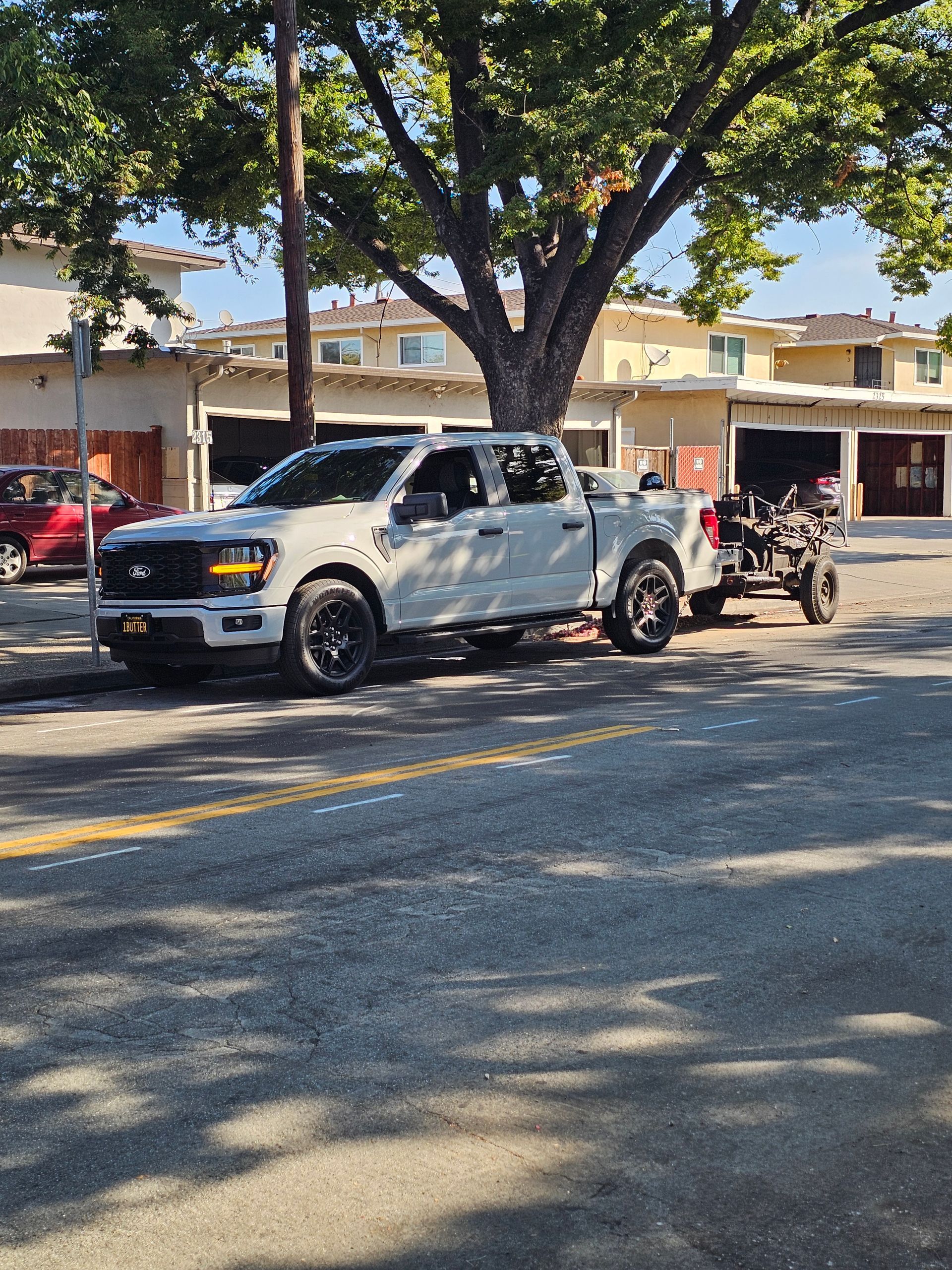 A white pickup truck parked on a residential street with a small all-terrain vehicle on a trailer attached behind it.