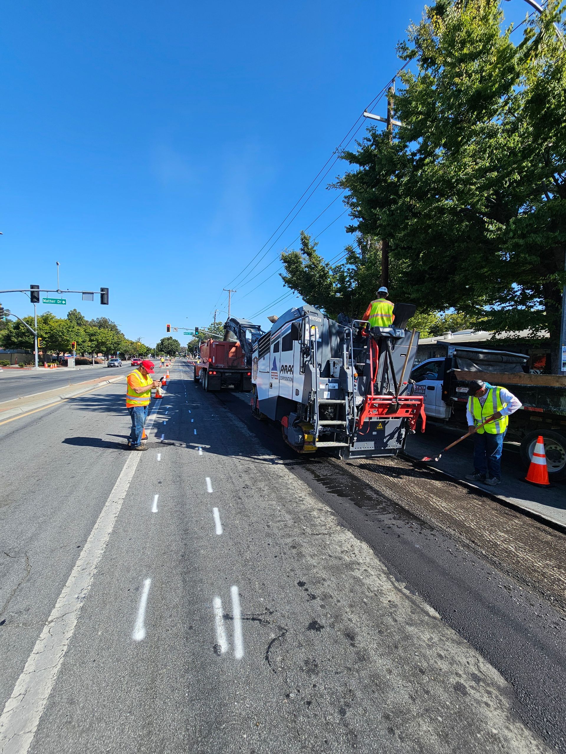 Road workers in high-visibility vests operate construction equipment on a sunny day next to a road with white lane markings.