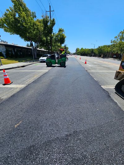 A small green road roller works on a newly paved dark asphalt strip on a sunny street lined with trees and safety cones.