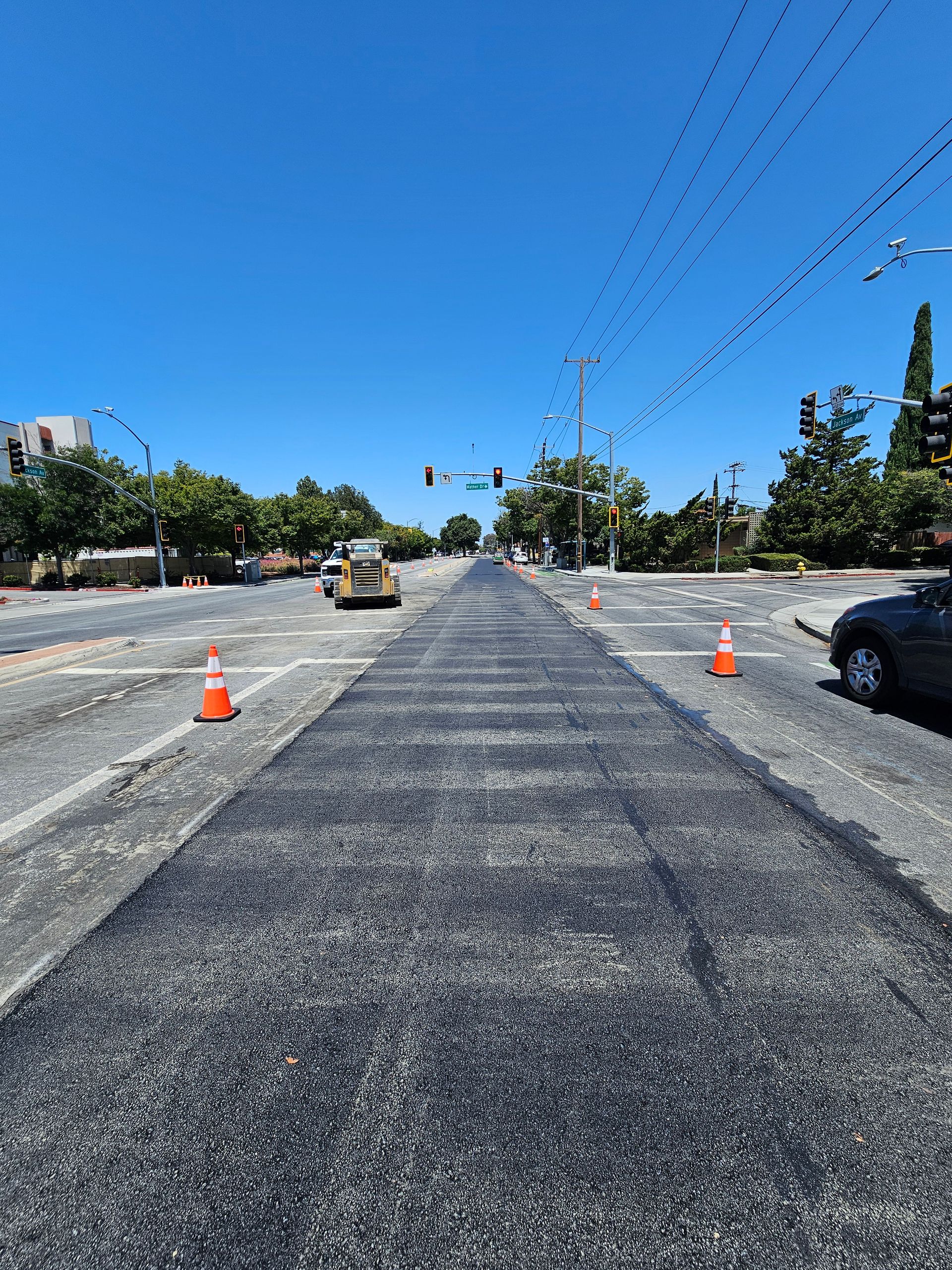 A freshly paved dark asphalt strip runs through the center of a sunlit street under construction with orange traffic cones.