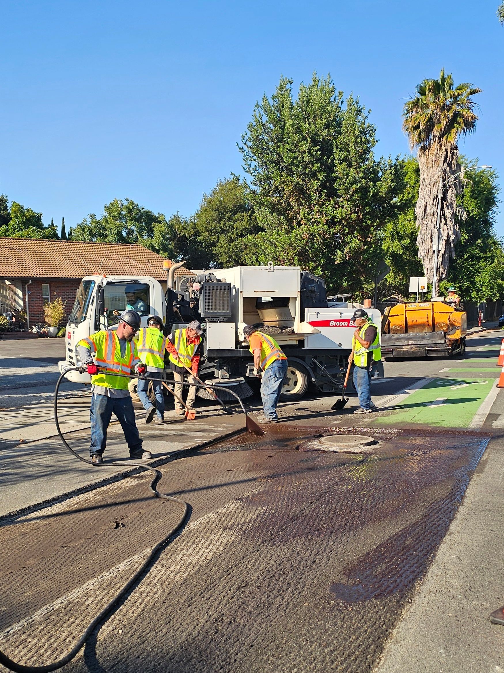 Construction workers in high-visibility vests work on a street repair site with specialized trucks and fresh asphalt.