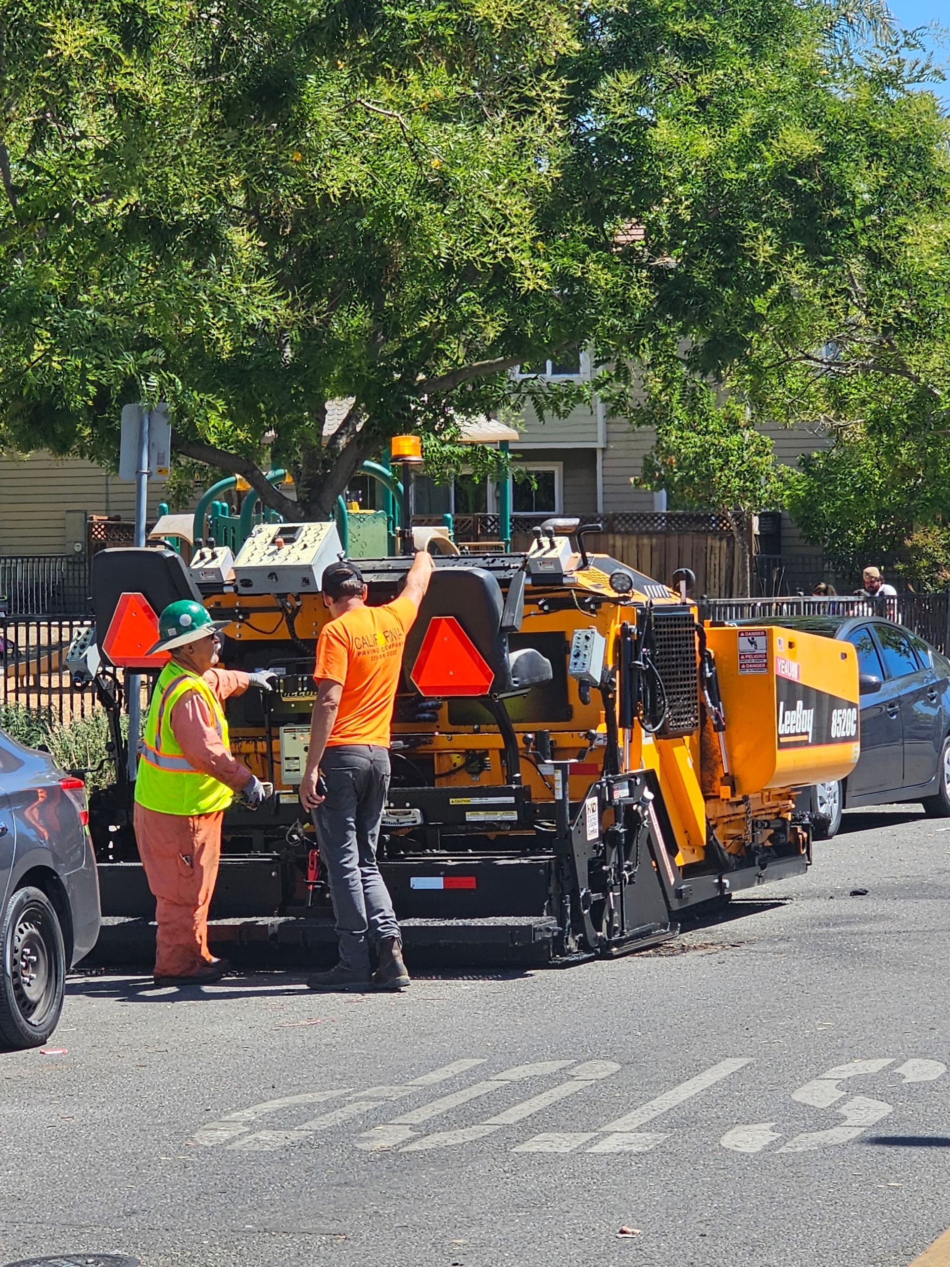 Two construction workers in high-visibility gear work on a yellow asphalt paving machine parked on a gravel road.